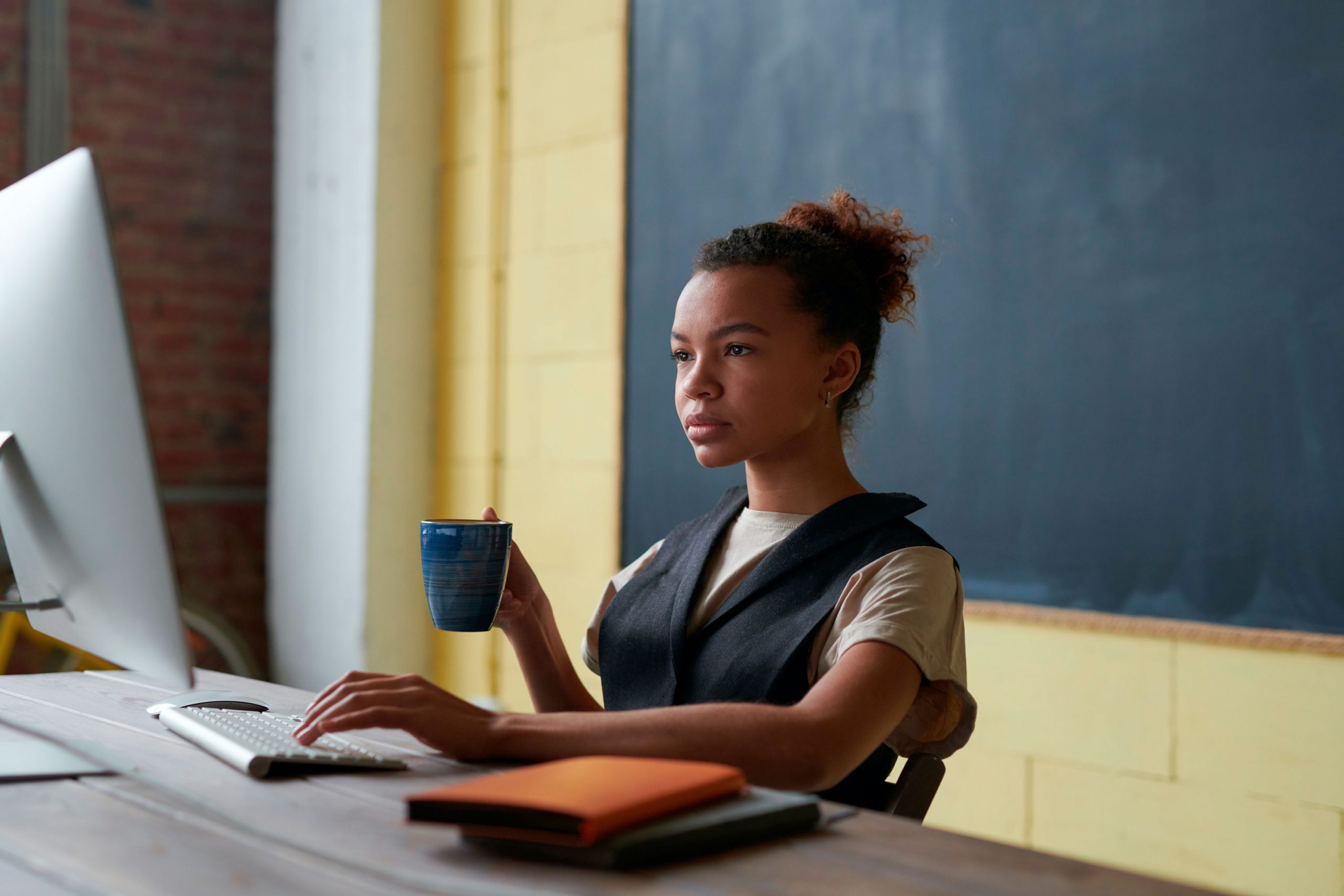 Uma mulher de pele negra, cabelos encaracolados presos em um coque, está sentada em frente a um computador, segurando uma xícara em uma mão e escrevendo no teclado com a outra. Sobre a mesa há diversos cadernos coloridos e atrás dela uma lousa